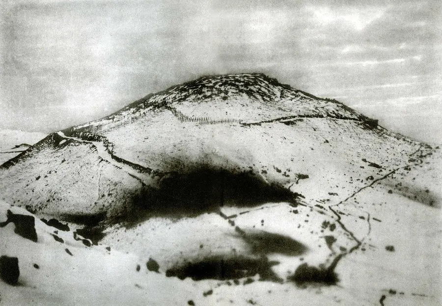 A snow-covered conical hill crossed by zigzag trench lines and pitted with shell craters