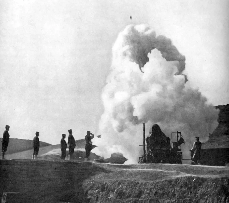 A heavy howitzer in a pit firing, a towering cloud of smoke, the shell a dot high above, gunners standing on the parapet